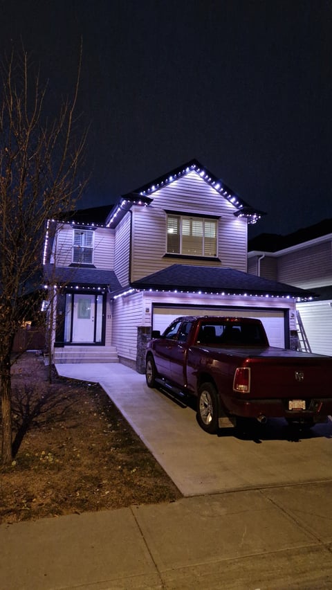 Two-story house with purple LED lights outlining the roofline and trim, burgundy pickup truck parked in driveway at night