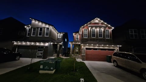 Two illuminated suburban houses at night with decorated rooflines, garage doors, and vehicles parked on the driveway