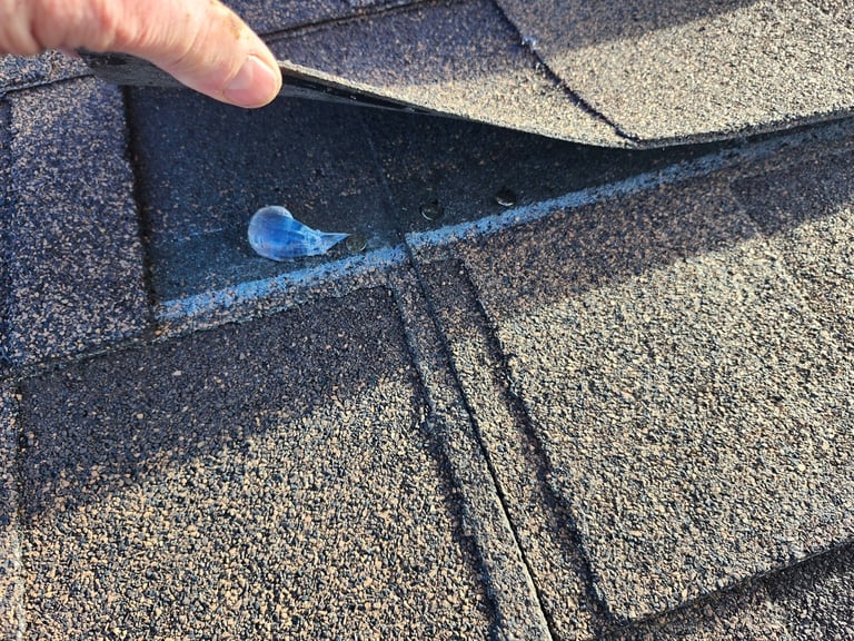 Hand lifting corner of asphalt shingles on a roof to reveal a small blue stone or granule beneath